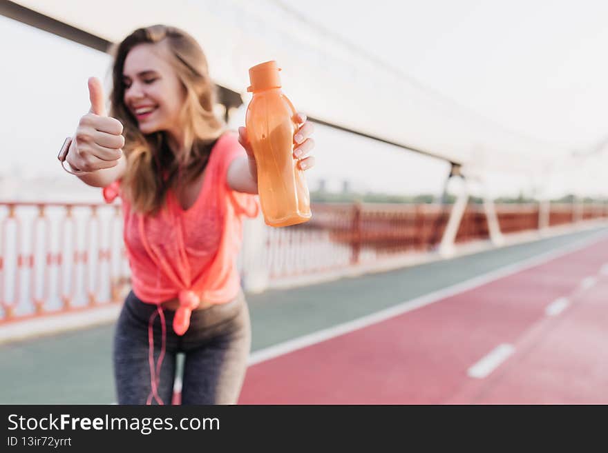 Refined girl in gray sport pants posing with smile. Laughing graceful woman standing with bottle at stadium.