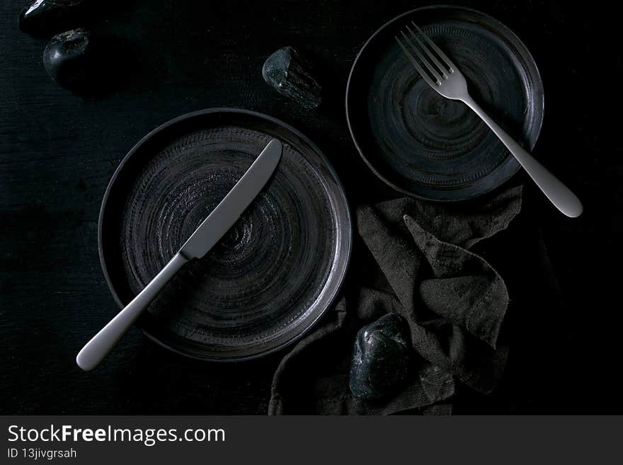 Empty black ceramic plates with cutlery and black stones around on textile napkin over black wooden background. Flat lay, space. Empty black ceramic plates with cutlery and black stones around on textile napkin over black wooden background. Flat lay, space.