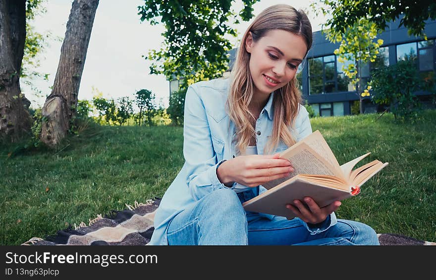 cheerful student reading book while sitting on checkered blanket in park,stock image