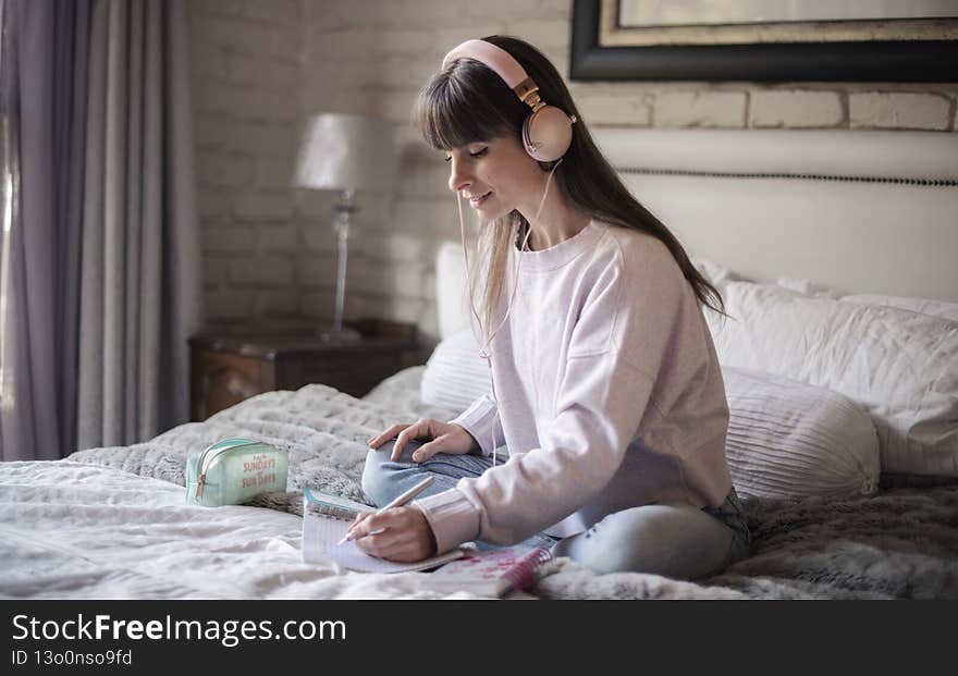 Young Woman on her bed in the Morning with her planner and pen writing notes into her planner whilst listening to music on her pink headphones.
