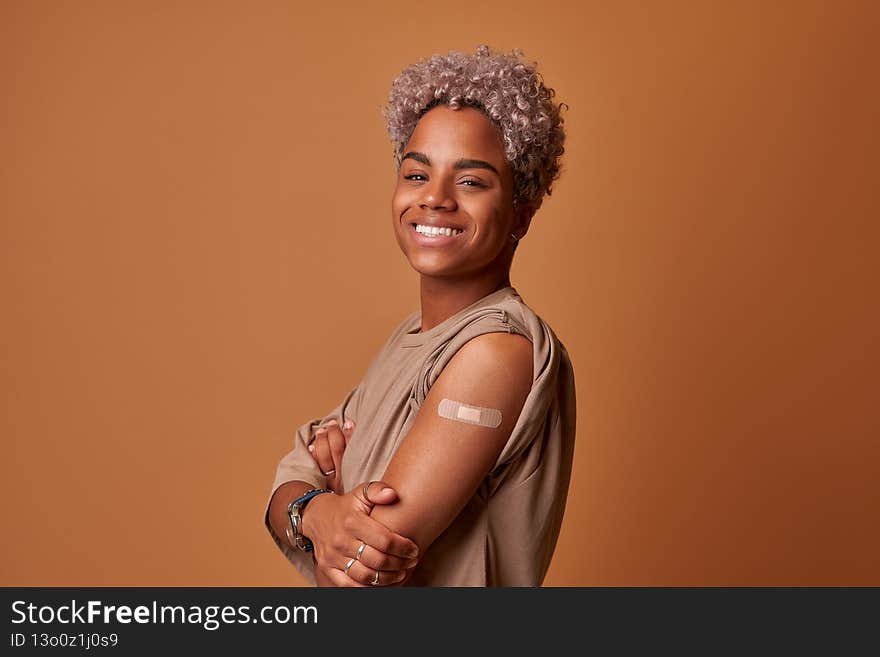 Happy Vaccinated Dark Skinned Woman Showing Arm With Plaster Bandage After Covid 19 Vaccine Injection Posing Over Champagne Background, Smiling To Camera. Coronavirus Vaccination Advertisement. Happy Vaccinated Dark Skinned Woman Showing Arm With Plaster Bandage After Covid 19 Vaccine Injection Posing Over Champagne Background, Smiling To Camera. Coronavirus Vaccination Advertisement.