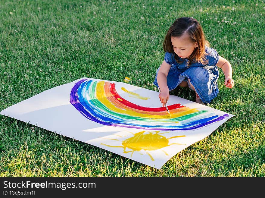 Little girl 2-4 years old paints rainbow and sun on large sheet of paper, sitting on green lawn
