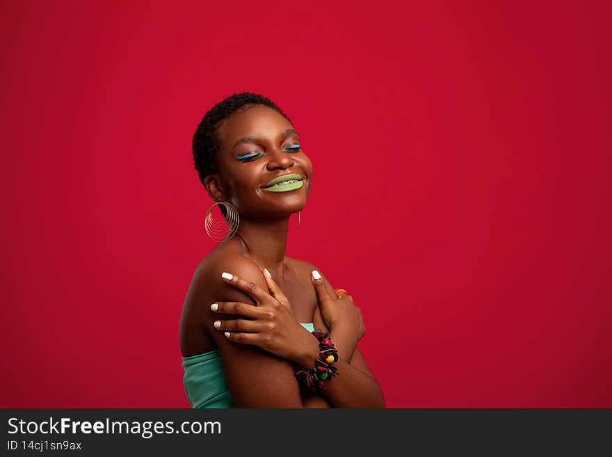 Exotic afro american young woman posing on red studio background, black female model wearing bright outfit and funky makep, using stylish accessories, hugging her shoulders and smiling, copy space. Exotic afro american young woman posing on red studio background, black female model wearing bright outfit and funky makep, using stylish accessories, hugging her shoulders and smiling, copy space