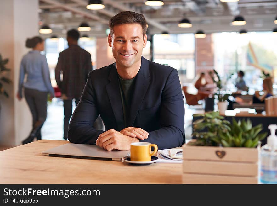 Portrait Of Smiling Businessman Sitting At Desk With Laptop In Modern Open Plan Office
