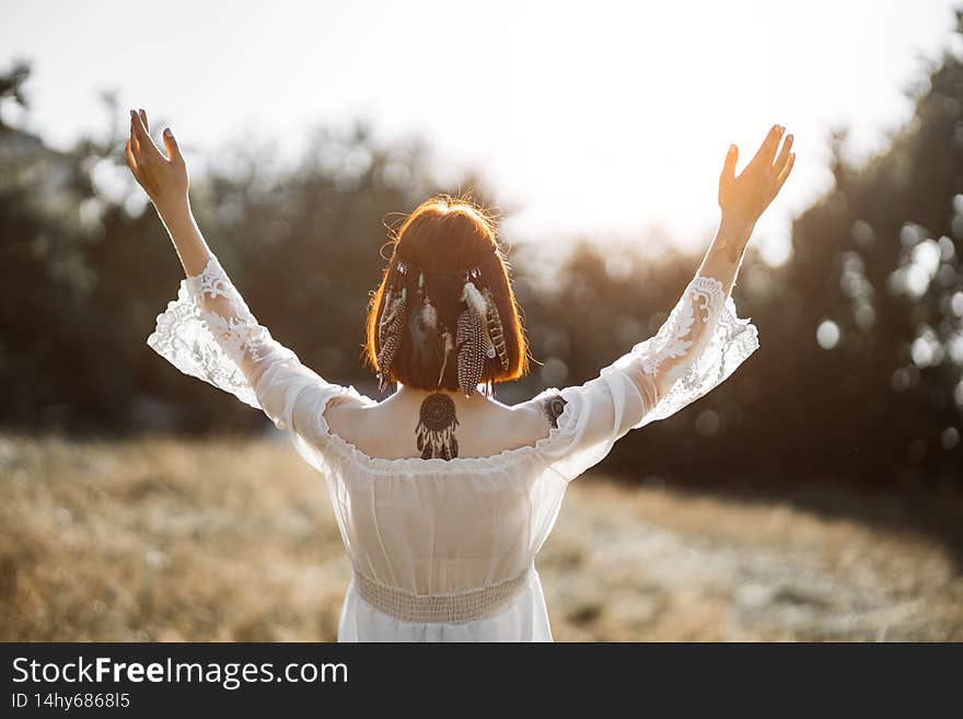 Indian boho vibes, bohemian style. Back view of stunning hippie boho woman with tattoos, wearing feathers in short ginger hair, white dress, stands with arms raised and outstretched to sunset in field