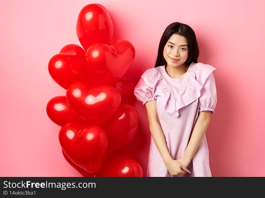 Cute asian girl in dress looking shy and smiling, standing modest near valentines day balloons, blushing on romantic