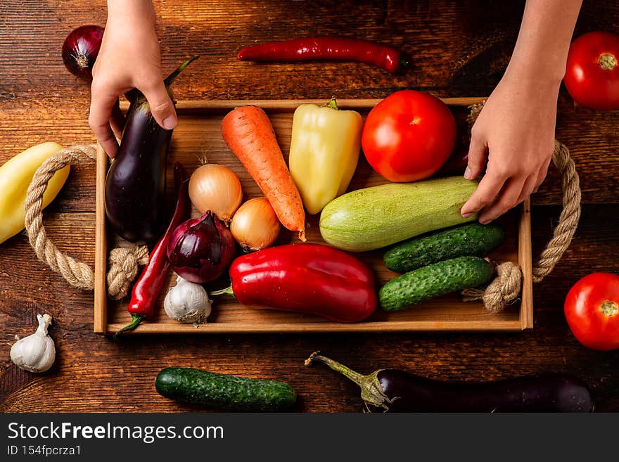 A woman holds a tray with different vegetables on a wooden background. Vegetable still life, background, vegetarianism concept