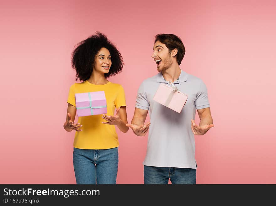 happy and thrilled interracial couple looking at each other near levitating gift boxes  on pink,stock image