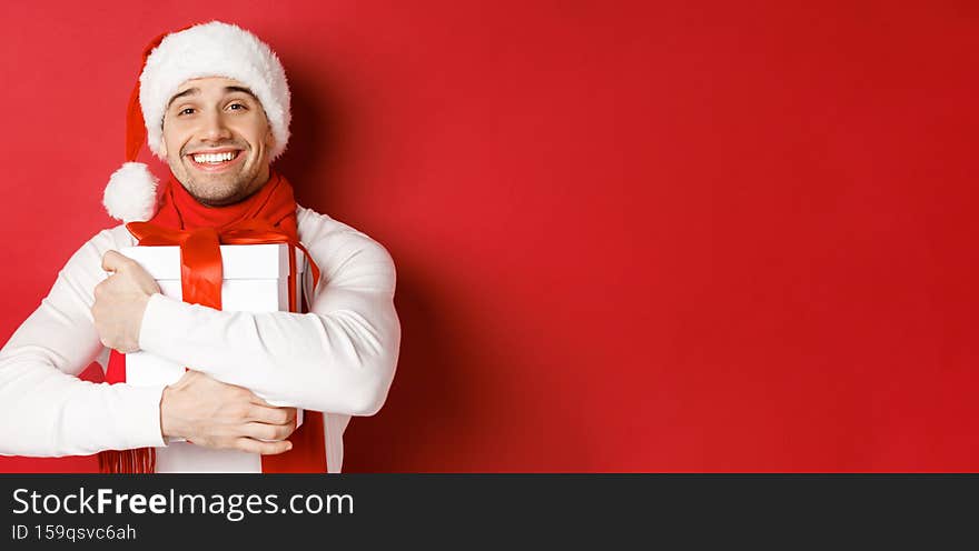 Concept of winter holidays, christmas and lifestyle. Image of lovely guy in santa hat and scarf, hugging his new year present and smiling flattered, standing over red background.