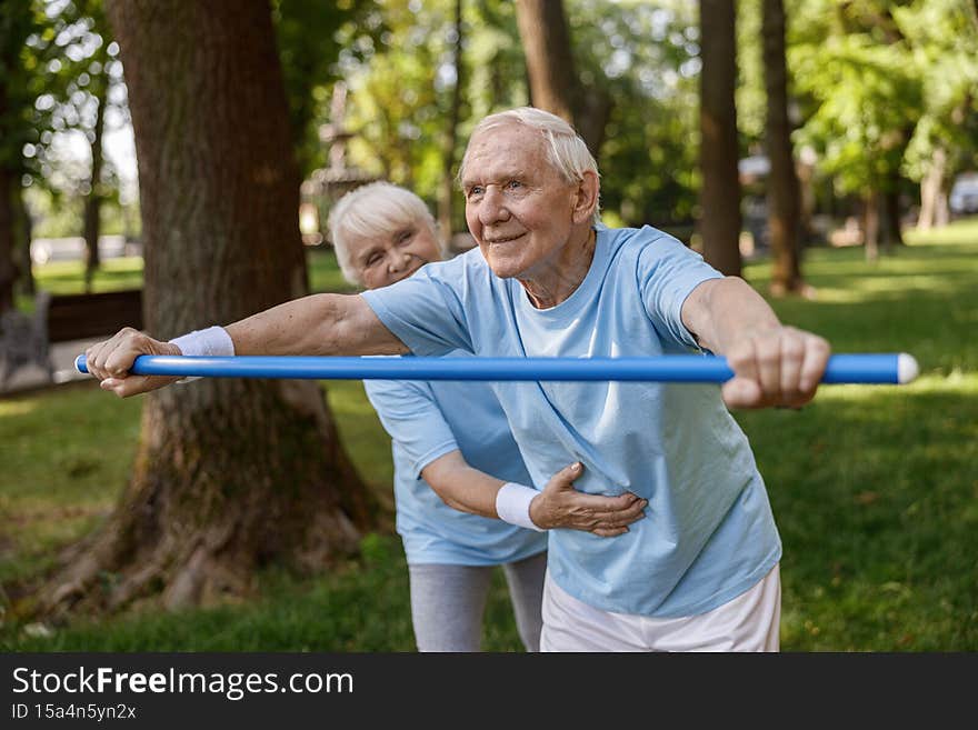 Senior gentleman does forward bend with bar rehabing injury with trainer help in green park on summer day. Active lifestyle and healthcare. Senior gentleman does forward bend with bar rehabing injury with trainer help in green park on summer day. Active lifestyle and healthcare
