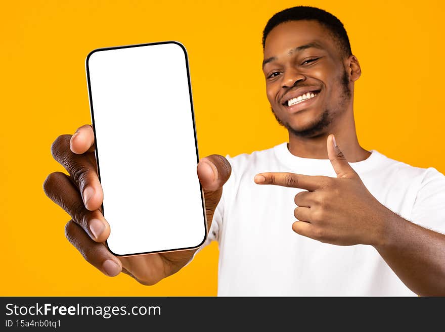 Cheerful handsome young black guy pointing at cellphone with blank screen and smiling at camera, mockup, recommending nice mobile application, yellow studio background, closeup photo. Cheerful handsome young black guy pointing at cellphone with blank screen and smiling at camera, mockup, recommending nice mobile application, yellow studio background, closeup photo