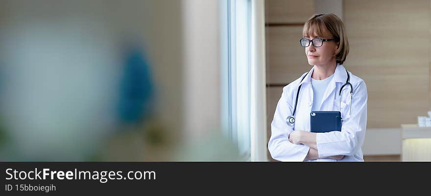 Caucasian senior woman doctor short blonde hair wearing medical uniform embracing tablet while standing near window and look out the window in the soft sunlight with Copy space in Banner size. Caucasian senior woman doctor short blonde hair wearing medical uniform embracing tablet while standing near window and look out the window in the soft sunlight with Copy space in Banner size