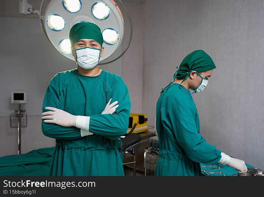 Doctors in operating room wearing green mask surgical gown surgeon hair cap latex glove one stand with arms crossed in front of bed and electrical defibrillator other while doctor prepare instruments