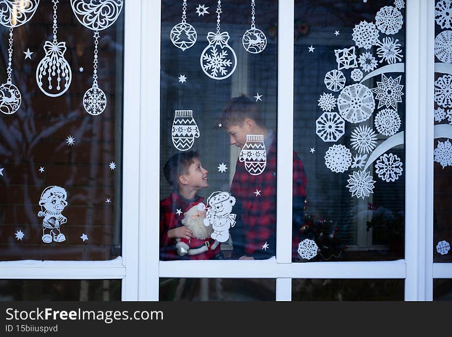 Handsome Kids standing by the decorated window with a toy santa claus and look and wait Christmas eve. Children hug and enjoy the winter holidays