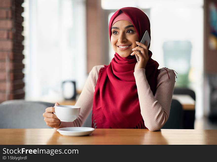 Happy young middle eastern lady in beautiful headscarf having phone conversation with girlfriend while drinking coffee, looking at copy space and smiling, enjoying coffee break, cafe interior. Happy young middle eastern lady in beautiful headscarf having phone conversation with girlfriend while drinking coffee, looking at copy space and smiling, enjoying coffee break, cafe interior