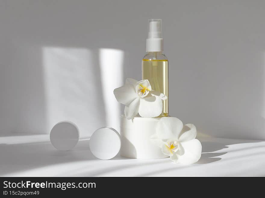 Mock-up of glass bottle with essential oil, two white jars with cream, surrounded by orchid flowers. White background with morning rays of sun. Concept of skin care, spa treatments.