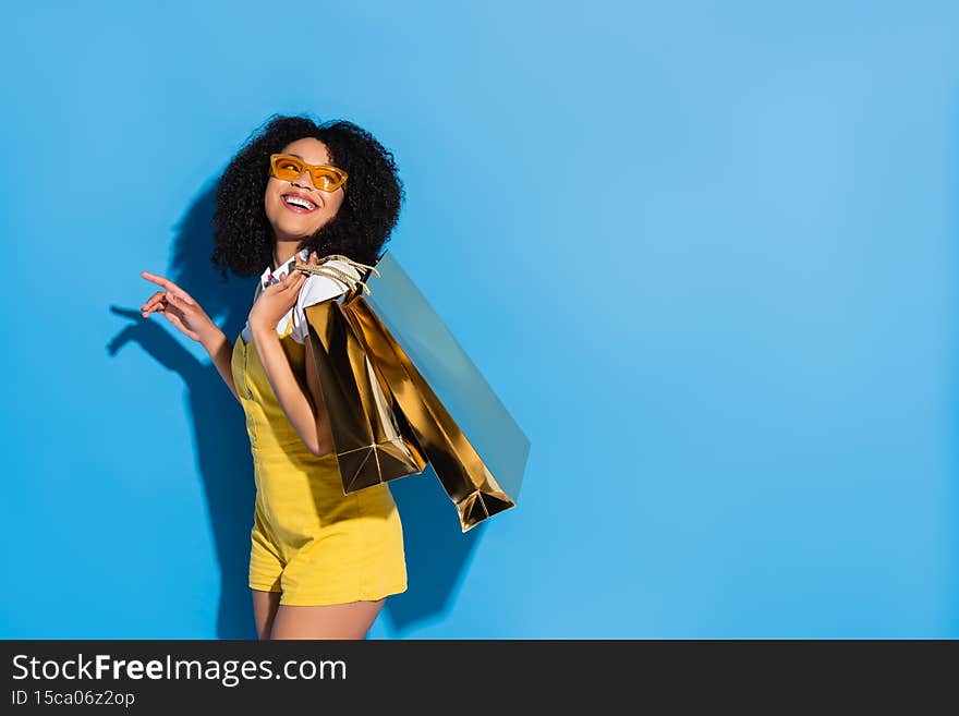 excited african american woman in yellow eyeglasses posing with shiny shopping bags on blue,stock image