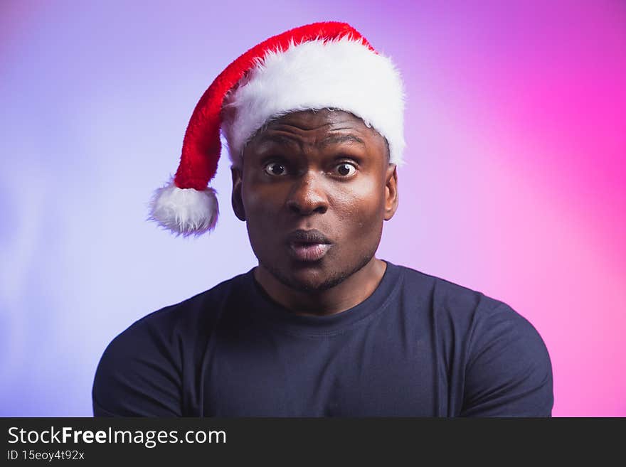 Portrait of surprised african american man in santa hat and casual t-shirt on colourful background, christmas time