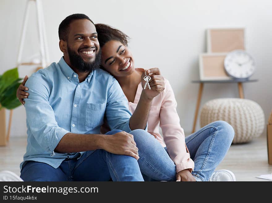 Glad millennial african american husband and wife moving to new apartment, sits among boxes and show house keys in new home. Insurance, real estate, mortgage, relocation, purchase flat and property