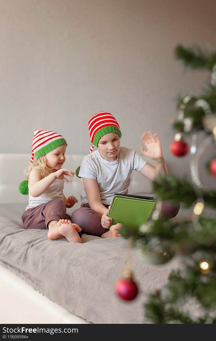 happy children brother and sister in pajamas and Christmas hats sitting home on bed and playing tablet, chatting online, greeting interlocutor. concept new normality, modern childhood