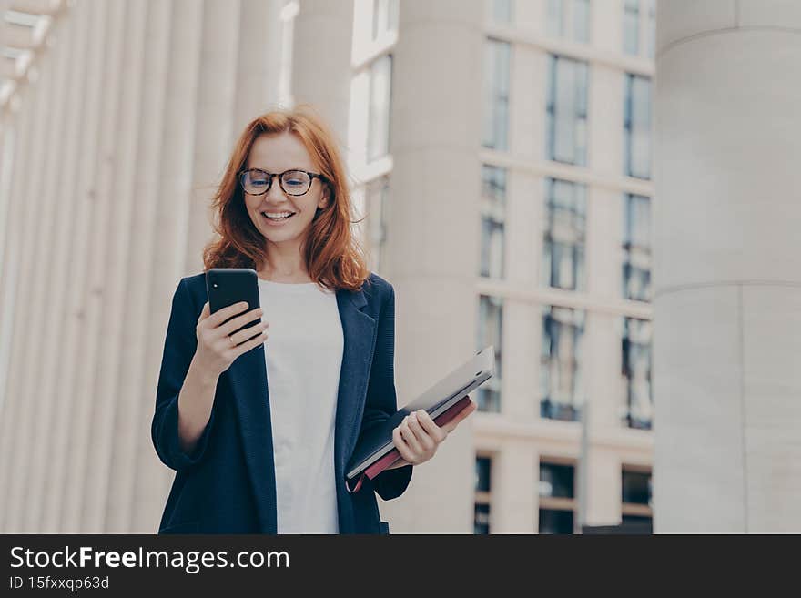 Young beautiful redhead female ceo executive looking at smartphone with excited face expression, reading news, browsing internet while going at business meeting with client, standing on city street