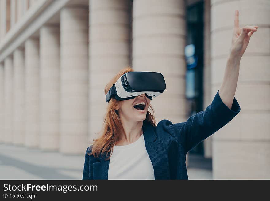 Amazed redhead businesswoman with opened mouth using VR glasses outside