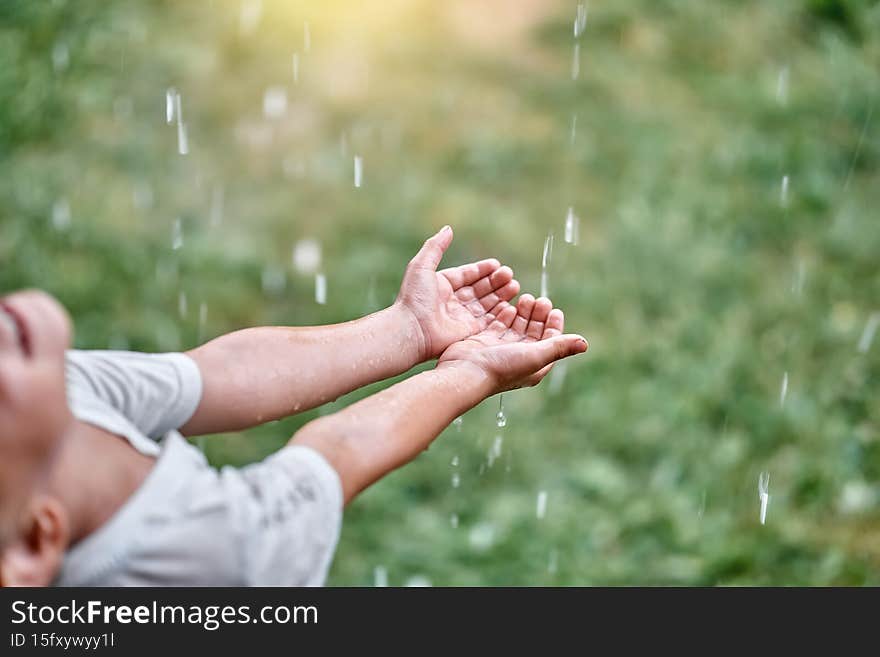 Cute little child boy enjoys warm summer rain catching falling drops with palms on green meadow in cottage backyard closeup. Cute little child boy enjoys warm summer rain catching falling drops with palms on green meadow in cottage backyard closeup