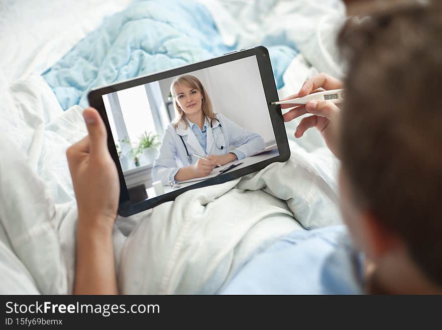 Telemedicine and online consultation concept. A young man or teenager sits in bed with an electronic thermometer and a