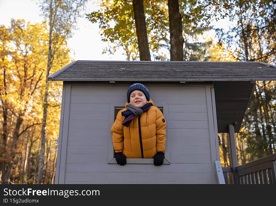 A cute smiling little boy peeks out of the window of a childrens house in the autumn on the playground. Concept childhood, nature. A cute smiling little boy peeks out of the window of a childrens house in the autumn on the playground. Concept childhood, nature.