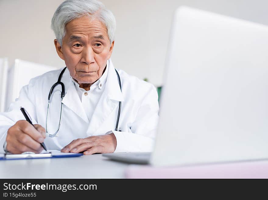 Elderly asian doctor writing on clipboard near blurred laptop in hospital,stock image