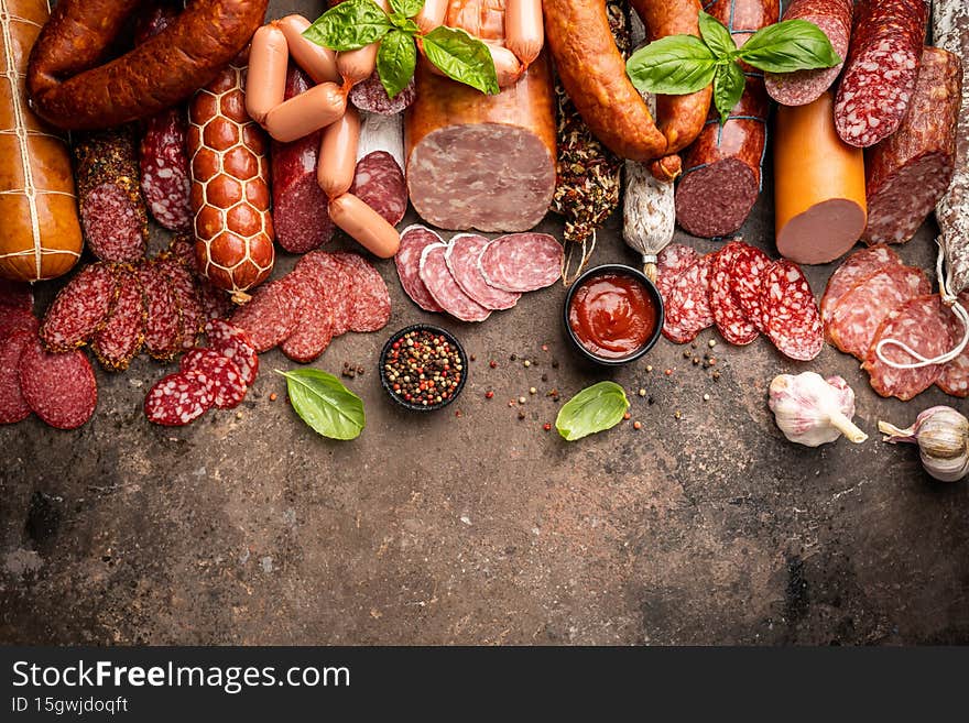 Set of different types of sausages, salami and smoked meat with basil and spices on brown stone background. Top view.