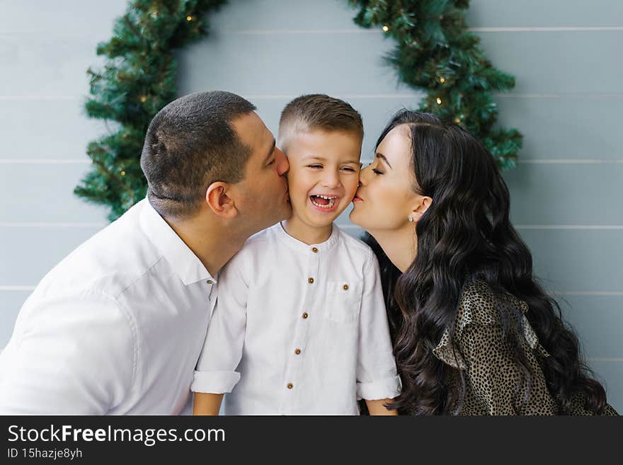 Beautiful young mom and dad kiss a cute little son standing against the background of a Christmas wreath on the wall.