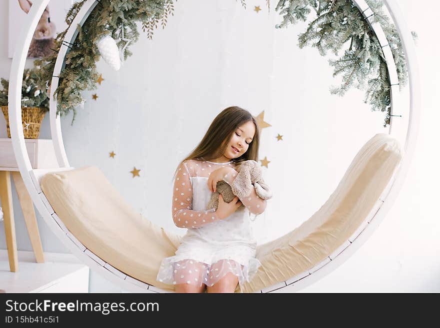 A small child smiles happily at the gift. A little girl with a cute rabbit on a Christmas swing in the children`s room. Little