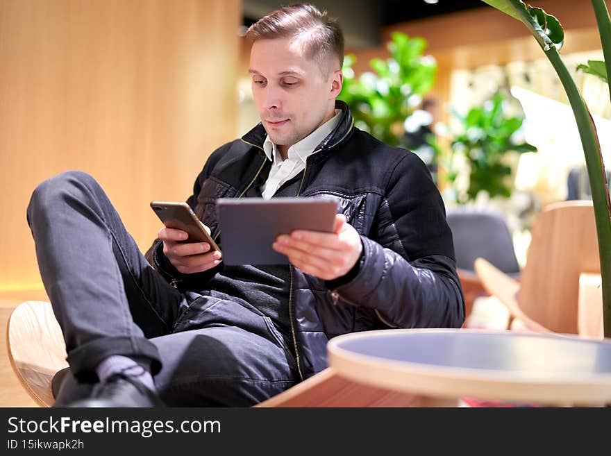 Male holding phone and tablet in his hands sitting on chair in store against green plant