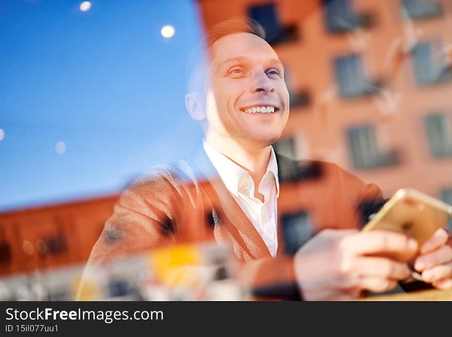 Happy male with phone in his hand sitting in cafe, building reflection in glass