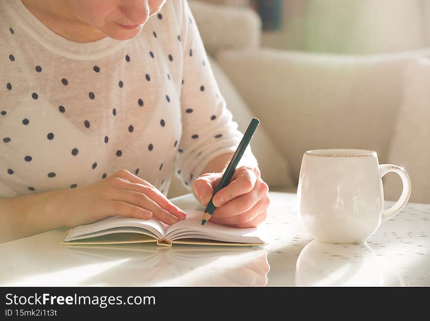 Woman sitting at the table, writing in the notebook and drinking coffee in nice light home interior. Working at home. Freelancer.