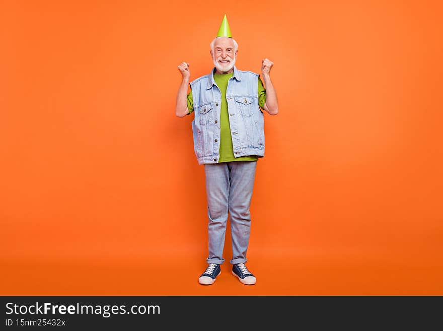 Photo of charming lucky retired man wear vintage jeans waistcoat birthday headwear rising fists smiling isolated orange