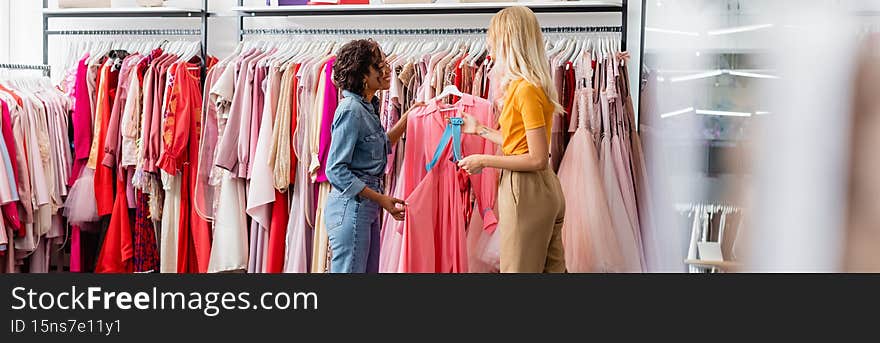 happy african american shop assistant showing pink dress to blonde and tattooed customer, banner,stock image
