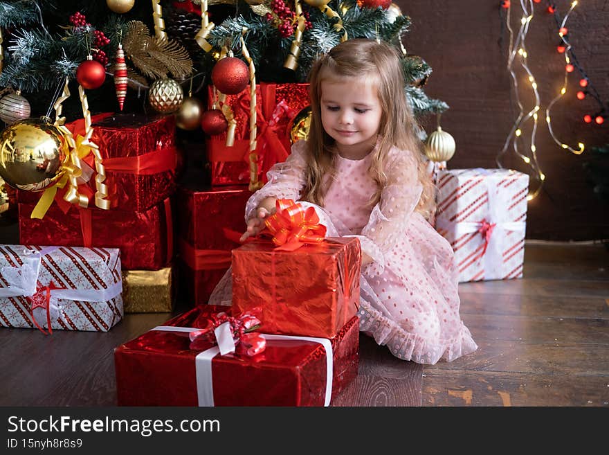 Happy little smiling blond girl in beautiful dress with gift box near christmas tree