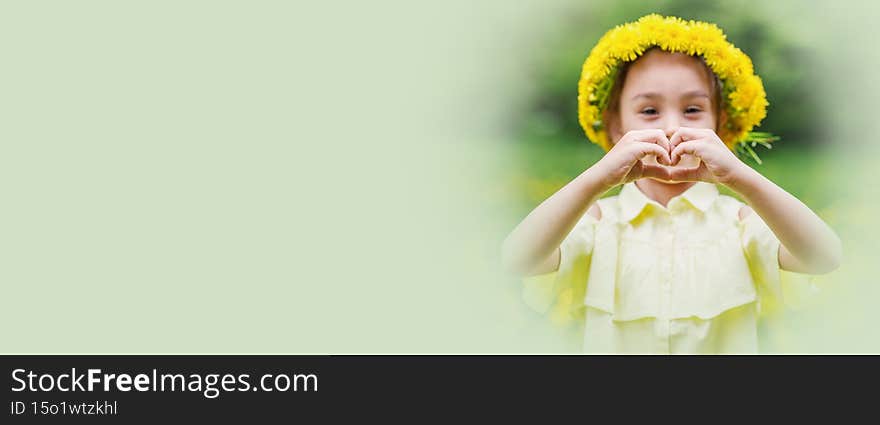 A girl in a wreath of dandelions shows a heart gesture with her hands. Walk in nature on a sunny summer day.