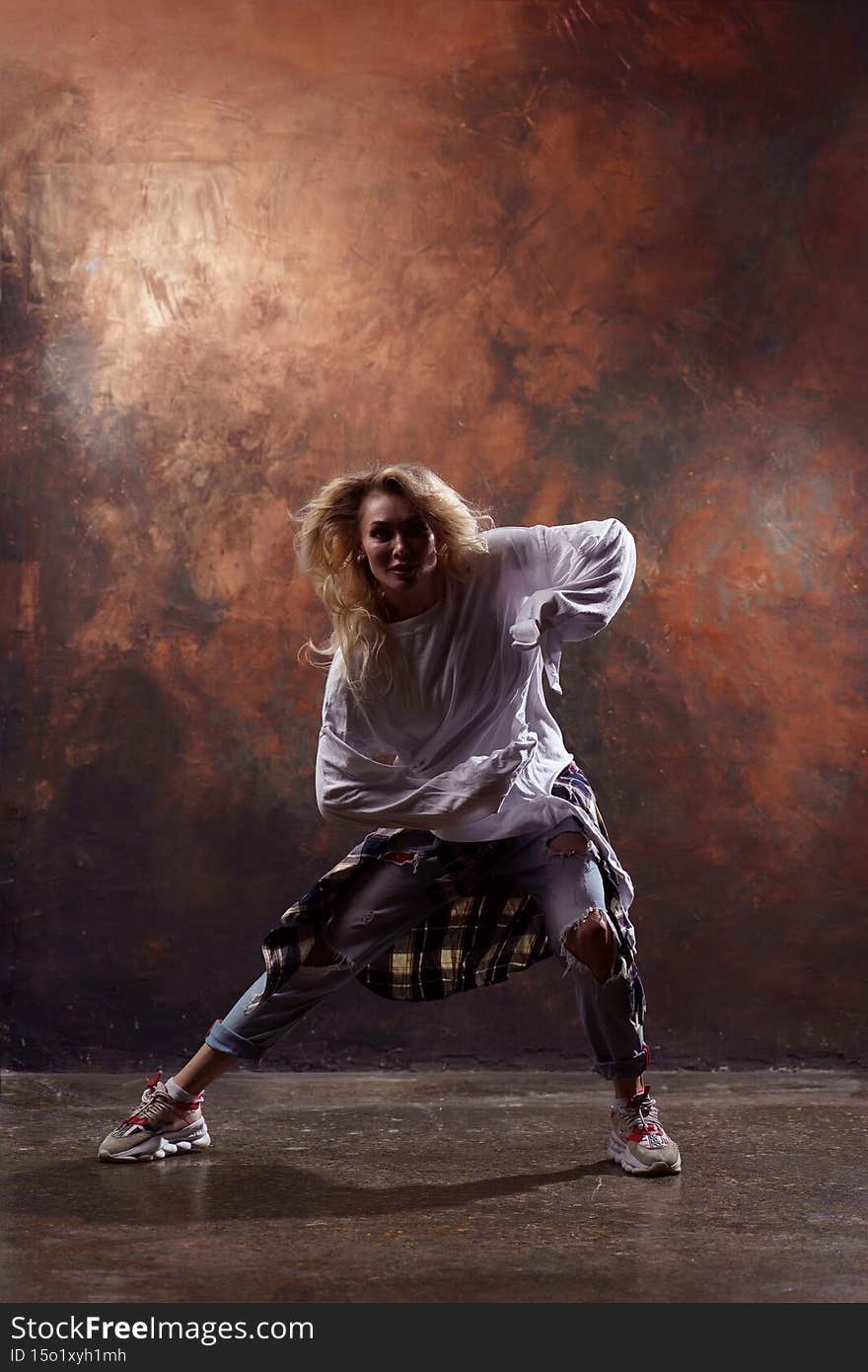 Photo of long-haired blonde dancer looking at camera in torn jeans and sneakers on dark brown background