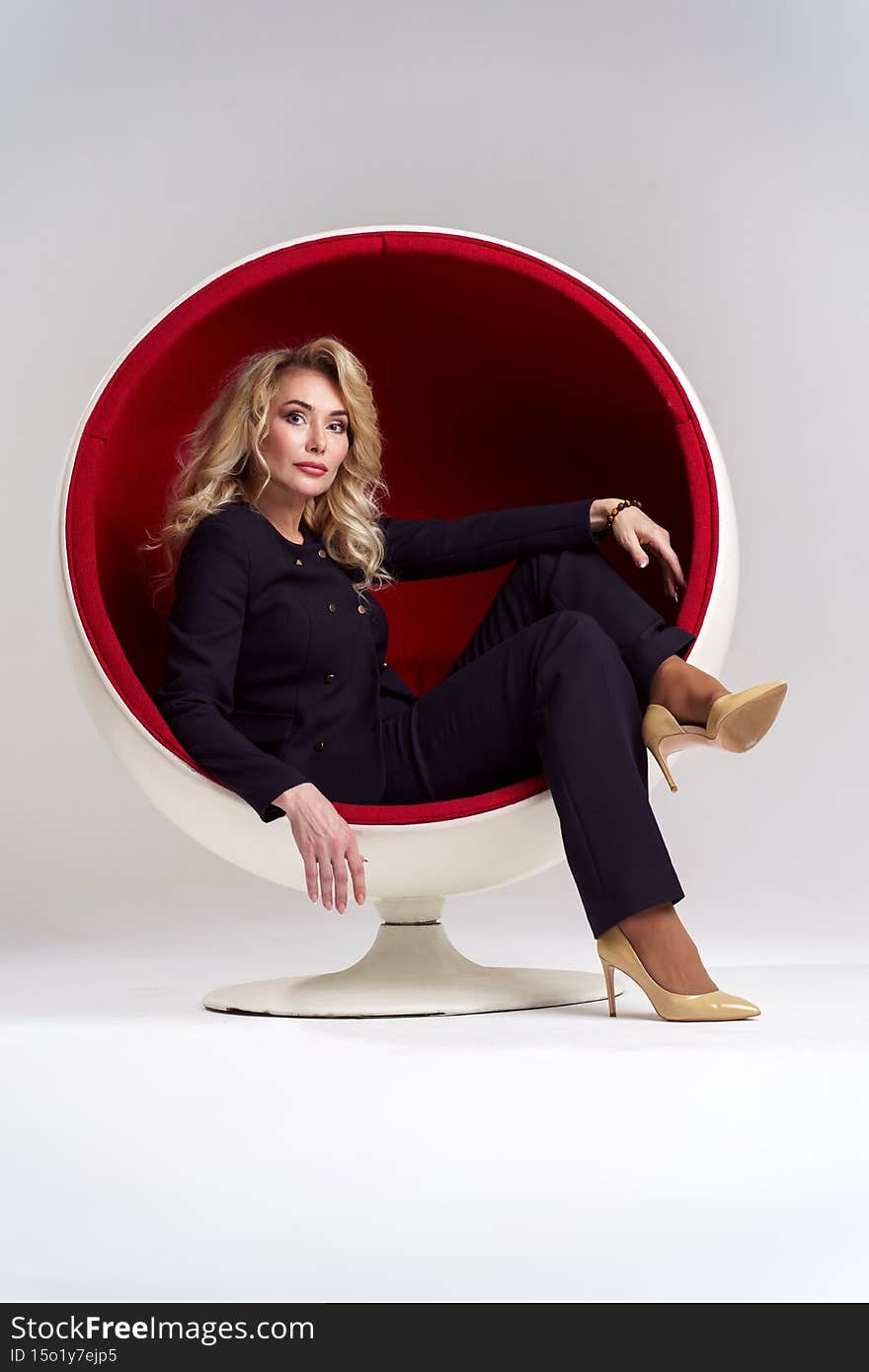 Image of blonde woman with long curly hair in black suit sitting in round chair  on white background in studio