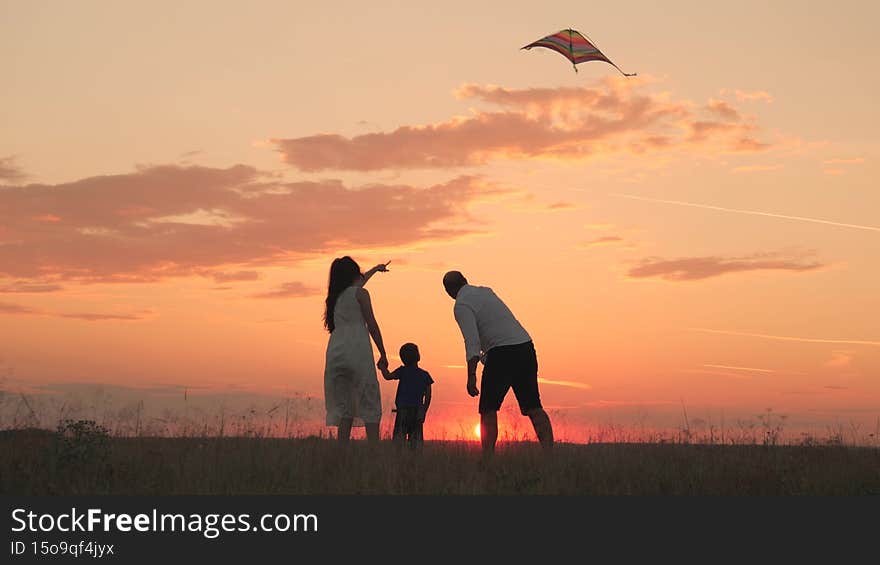 Happy family, little child with father and mother launch flying kite into sunset sky, silhouette of mom dad and kid