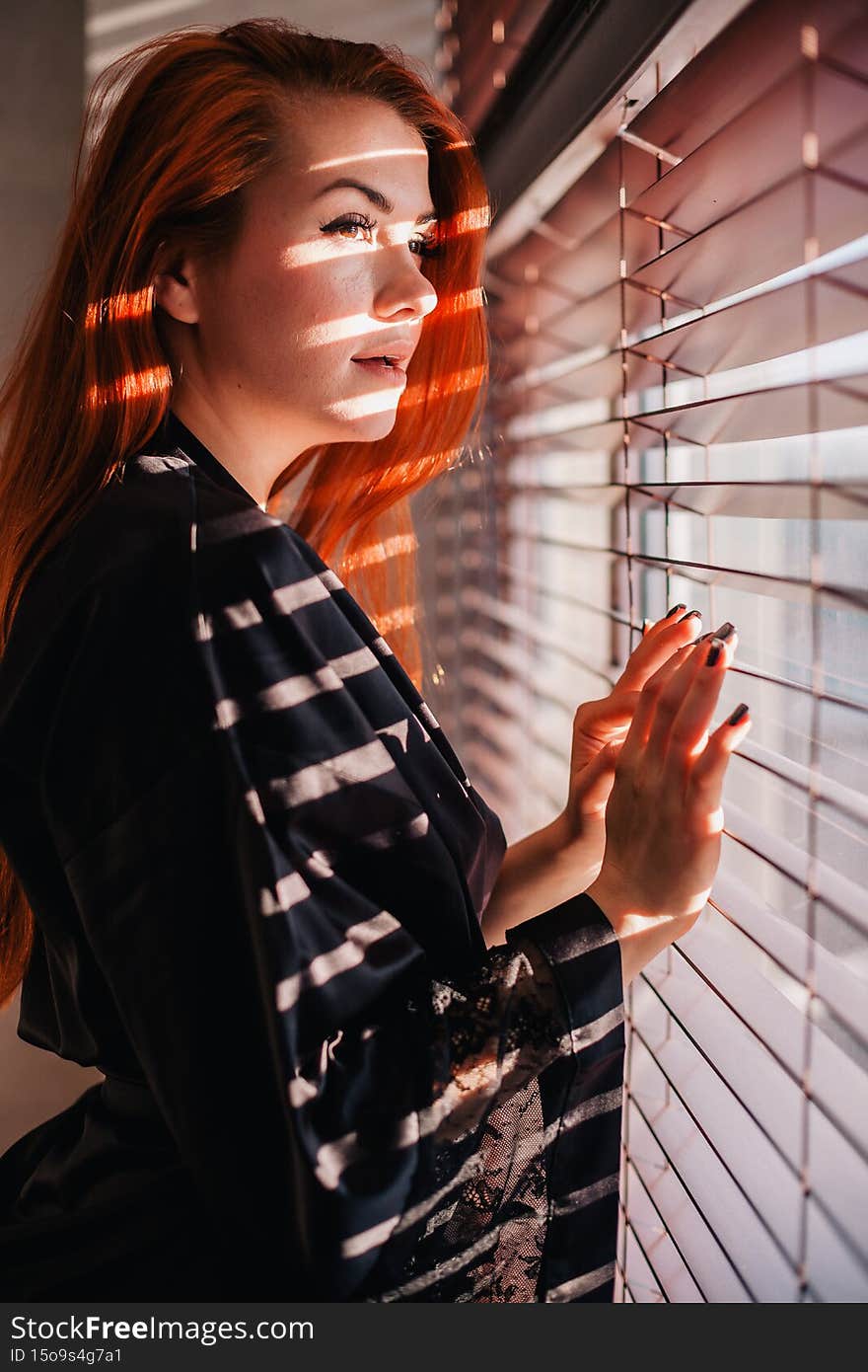 A red, slender girl in a beautiful black robe stands by the window in the light of the sun through the blinds.