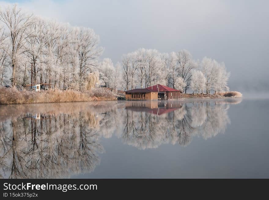 House by the lake. The transition of nature from autumn to winter. Foggy and sunny morning on the lake.