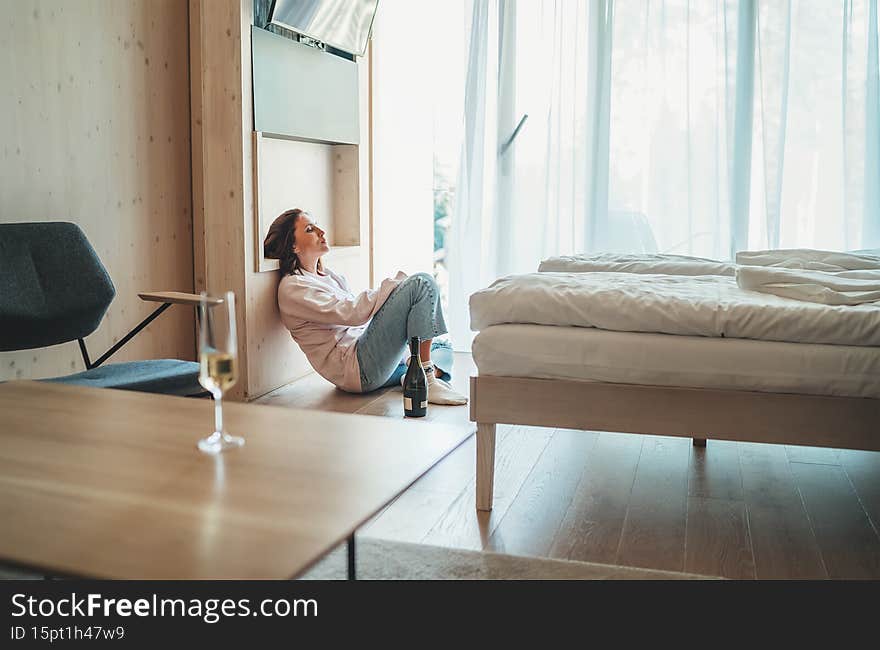 Sad woman sitting on the floor next to window in the bedroom with an opened bottle of alcohol. Unfocused white wine glass on the