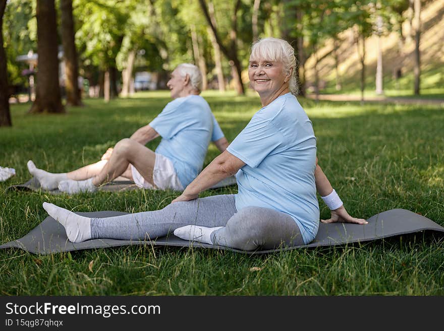 Positive mature woman with friend do yoga asanas on field grass in picturesque park