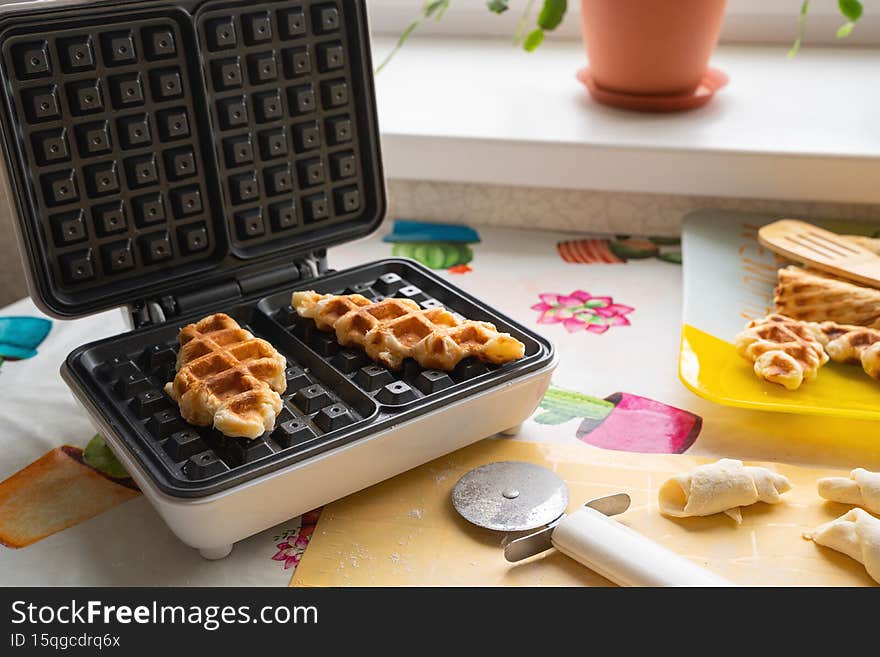 The croffle waffles are in a waffle iron and on a platter. Billets of puff-yeast dough are on the placemat. Culinary trend, Asian dessert. The croffle waffles are in a waffle iron and on a platter. Billets of puff-yeast dough are on the placemat. Culinary trend, Asian dessert..