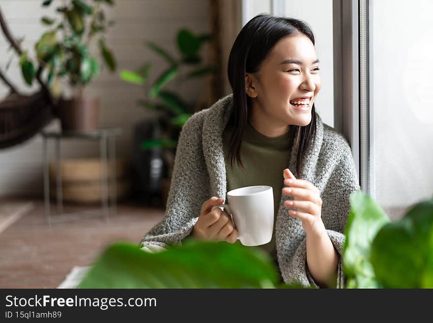 Happy asian woman drinking morning coffee and laughing, looking outsie window cheerful.