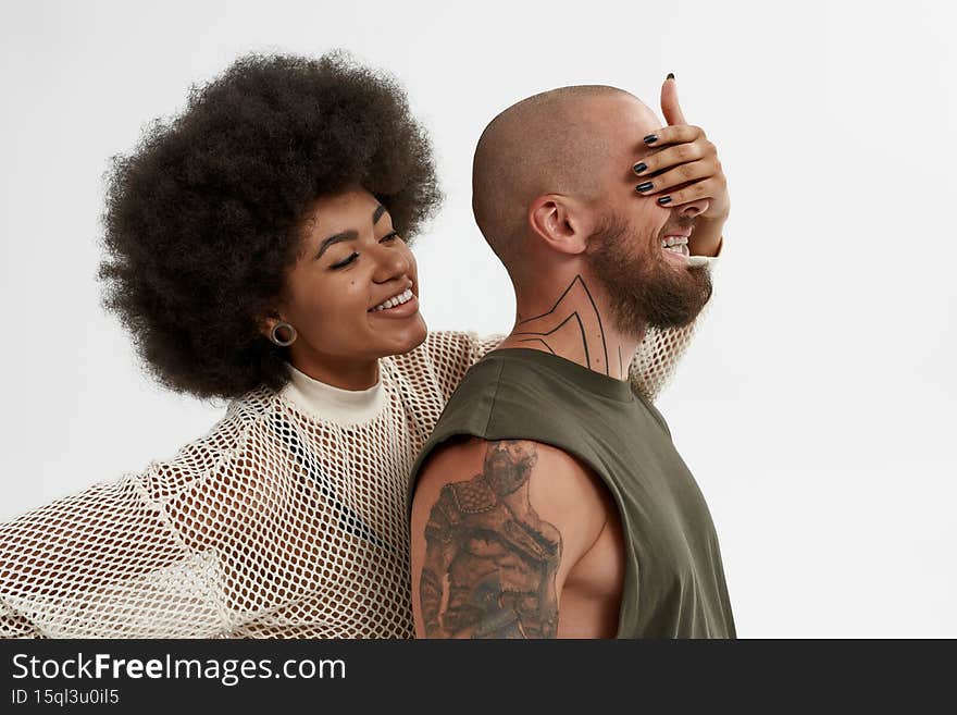 Black women covering eyes of her male friend. Concept of friendship. Side view of young smiling couple of bearded bald guy and curly brunette girl. Isolated on white background in studio. Black women covering eyes of her male friend. Concept of friendship. Side view of young smiling couple of bearded bald guy and curly brunette girl. Isolated on white background in studio